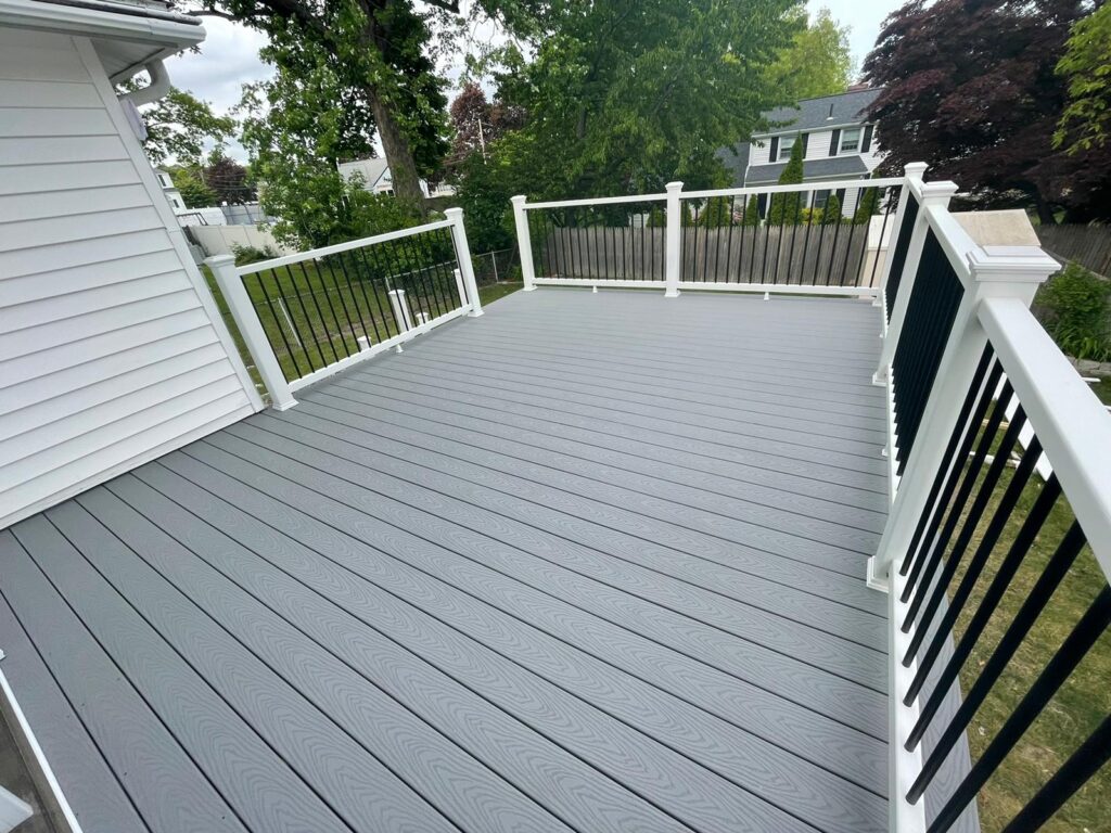 Full view of completed elevated gray composite deck in Quincy, MA by Monroy's Carpenters, Inc., attached to white-sided home with black railings, steps,
