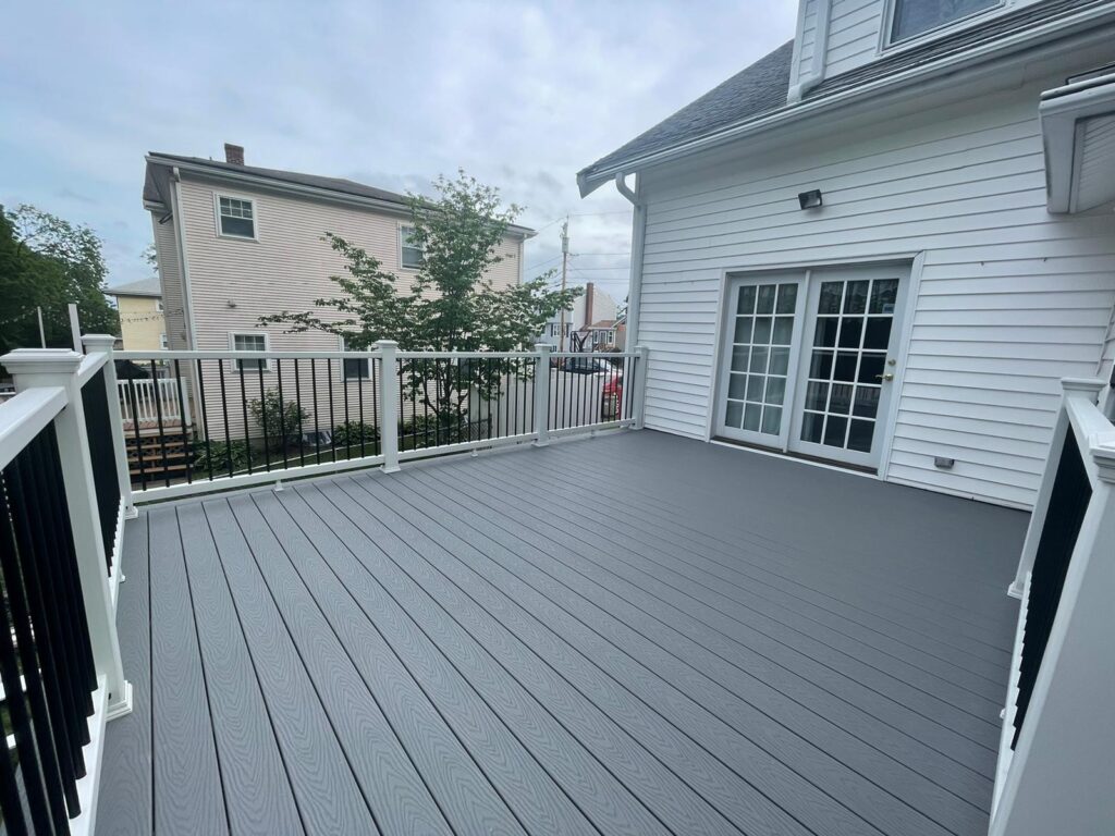 Spacious gray deck surface with black metal railings and white posts on elevated structure built by Monroy's Carpenters in Quincy, Massachusetts, overlooking green lawn.