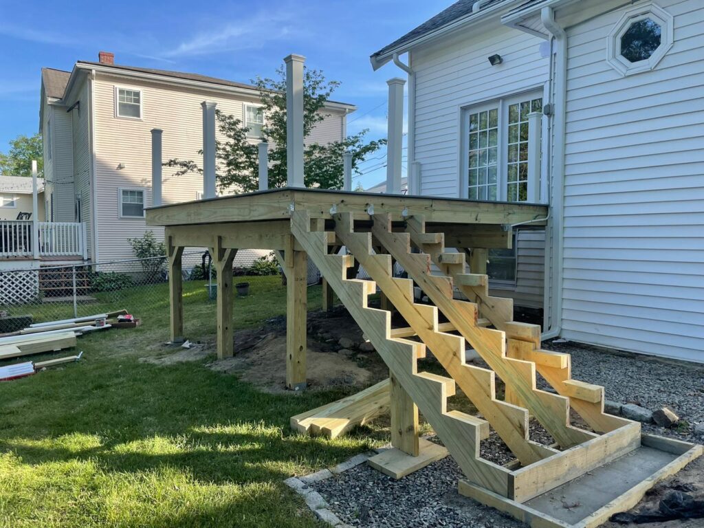 Progress photo of elevated deck framing and wooden steps during construction in Quincy, MA by Monroy's Carpenters, Inc., with lumber stacks and house integration visible.