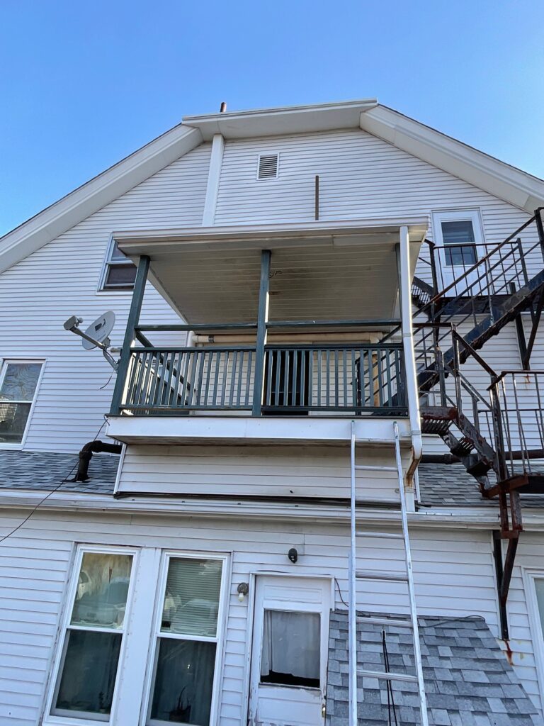 Rear view of a two-story house in Waltham, MA, featuring a deck under construction by Monroy's Carpenters, Inc. The deck has a green railing and a metal fire escape,