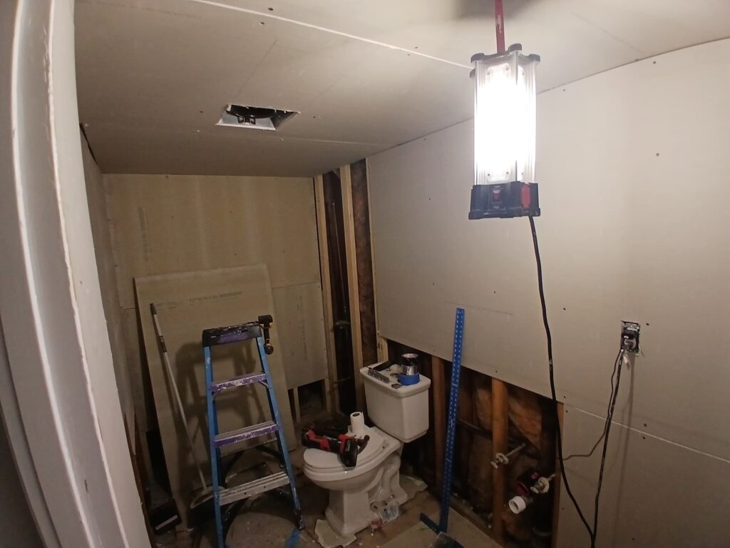 The "before" photo shows a bathroom in the midst of a renovation by Monroy's Carpenters, Inc. The space is under construction, with unfinished walls featuring exposed drywall and framing. A toilet is installed but surrounded by debris, tools, and a ladder, indicating active work. Plumbing pipes and electrical wiring are visible, and a bright work light illuminates the area, highlighting the incomplete state with a hole in the ceiling and scattered materials on the floor.