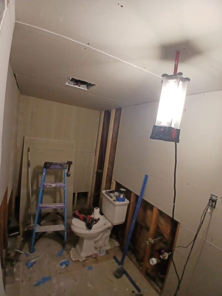 The "before" photo shows a bathroom in the midst of a renovation by Monroy's Carpenters, Inc. The space is under construction, with unfinished walls featuring exposed drywall and framing. A toilet is installed but surrounded by debris, tools, and a ladder, indicating active work. Plumbing pipes and electrical wiring are visible, and a bright work light illuminates the area, highlighting the incomplete state with a hole in the ceiling and scattered materials on the floor.