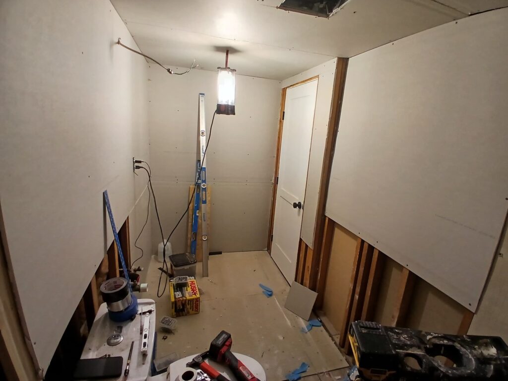 The "before" photo shows a bathroom in the midst of a renovation by Monroy's Carpenters, Inc. The space is under construction, with unfinished walls featuring exposed drywall and framing. A toilet is installed but surrounded by debris, tools, and a ladder, indicating active work. Plumbing pipes and electrical wiring are visible, and a bright work light illuminates the area, highlighting the incomplete state with a hole in the ceiling and scattered materials on the floor.