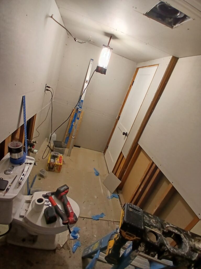 The space is under construction, with unfinished walls featuring exposed drywall and framing. A toilet is installed but surrounded by debris, tools, and a ladder, indicating active work.