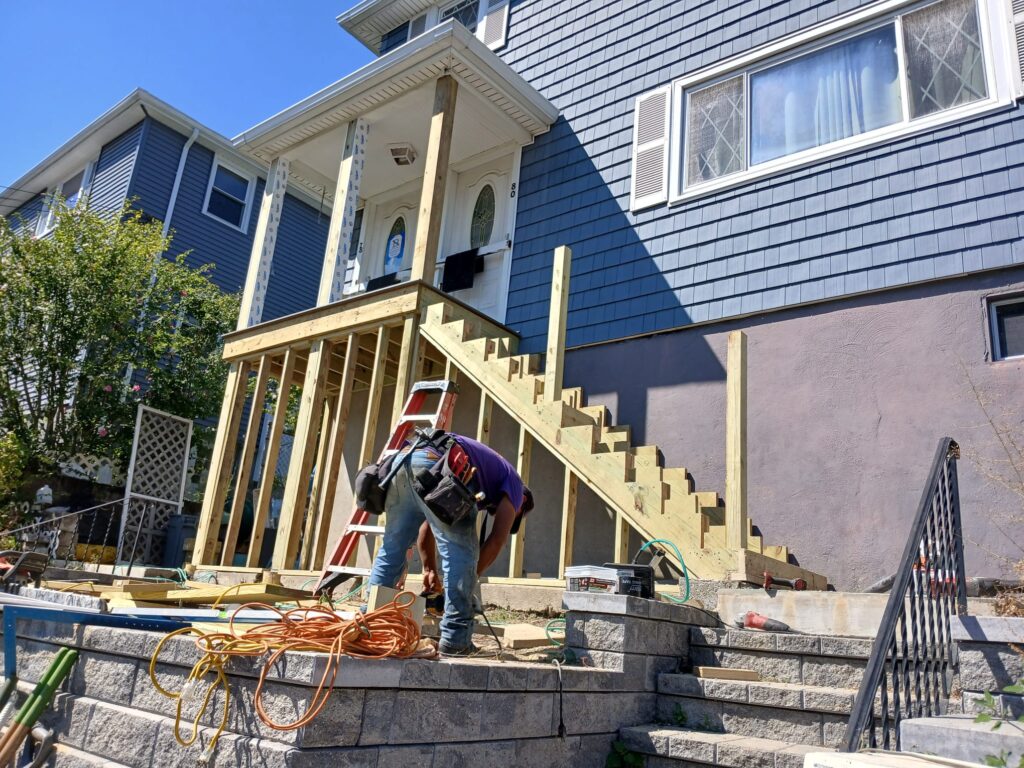 Lower angle of deck building by Monroy's Carpenters Inc in Brighton MA: Wooden stair frame and supports on a blue house, with a worker and construction materials, showcasing quality deck work in Boston suburbs