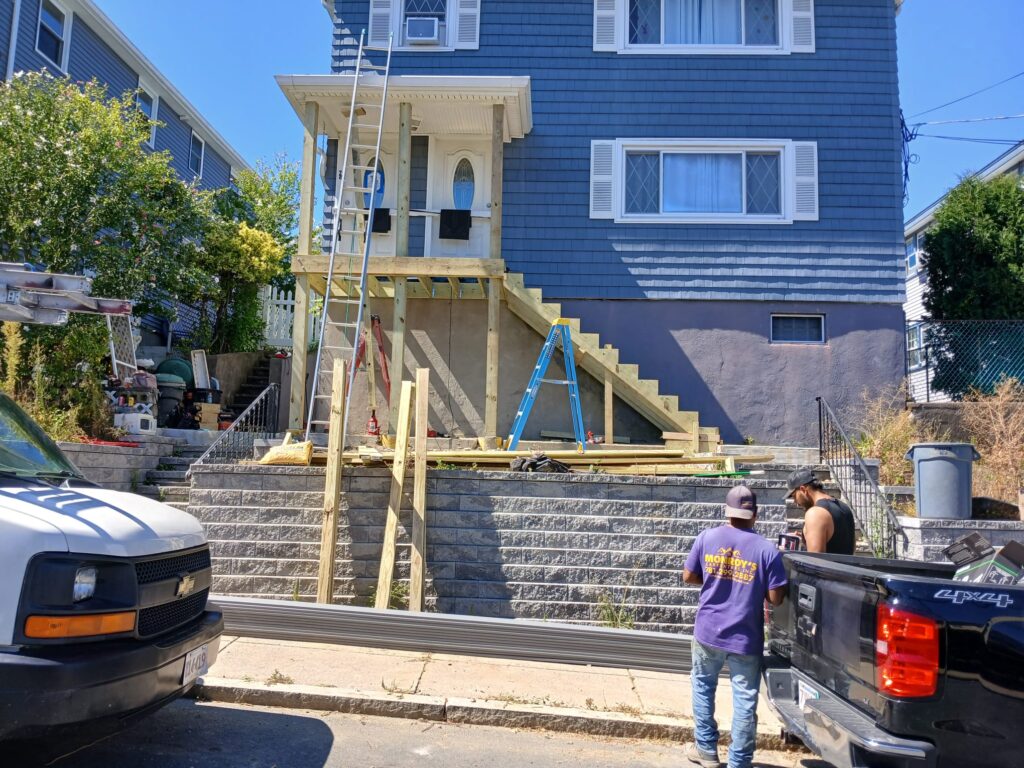 Custom porch stairs by Monroy's Carpenters Inc in Brighton MA: Wooden stair frame with a worker in a purple shirt and tool belt, near a blue house, demonstrating skilled outdoor home improvement in Boston.