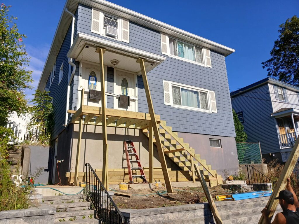 Front view of deck framework by Monroy's Carpenters Inc in Brighton MA: Wooden structure with columns on a blue house exterior, ladders, and tools, enhancing outdoor living spaces in Greater Boston.