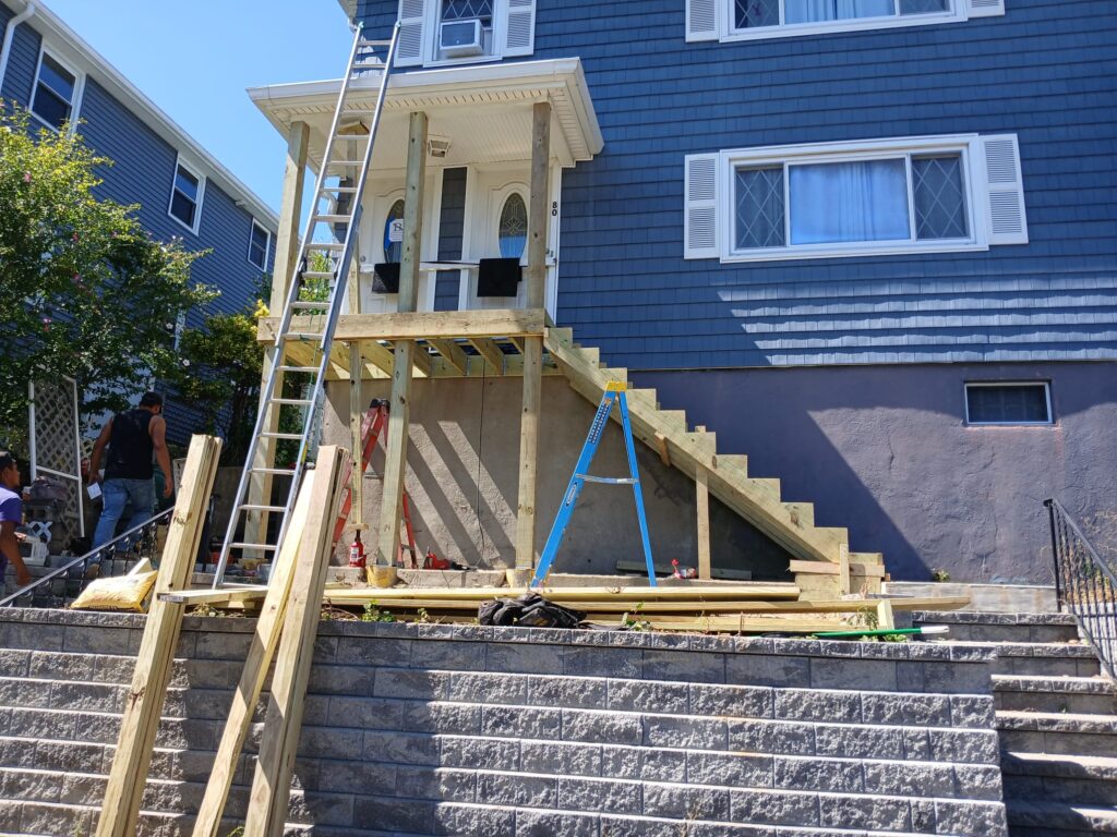 Porch framing by Monroy's Carpenters Inc in Brighton MA: Wooden frame structure with stairs under construction on a blue house, workers using ladders, showcasing expert deck building in Greater Boston.