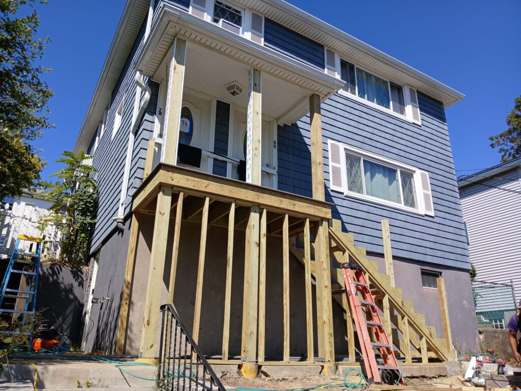 Monroy's Carpenters Inc porch construction in Brighton MA: Wooden supports and stairs being built on a blue-sided home, with tools and ladders, highlighting custom porch installation in Waltham area.