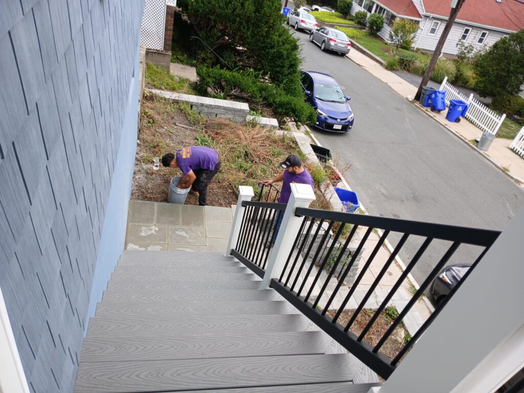 Monroy's Carpenters Inc deck stairs in Brighton MA: Gray composite stairs with black metal railings, a worker in a purple shirt adding finishing touches, showcasing expert carpentry for durable outdoor spaces in Waltham.