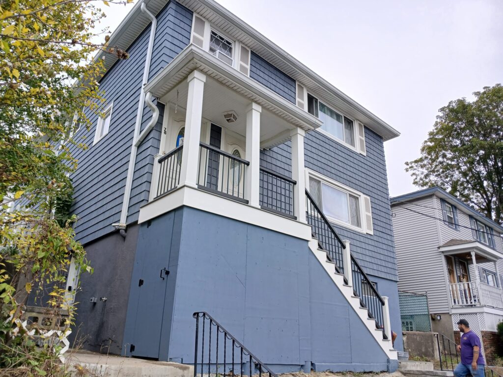 Two-story residential house with blue-gray siding, white trim, and a newly renovated elevated porch entrance. A black metal railing with white posts borders the staircase leading up to the front door. The porch has decorative glass double doors, square columns, and a white roof overhang. Below the porch, a blue-painted wall and storage access doors are visible. A man is walking on the sidewalk in front of the property, and neighboring houses are seen in the background.