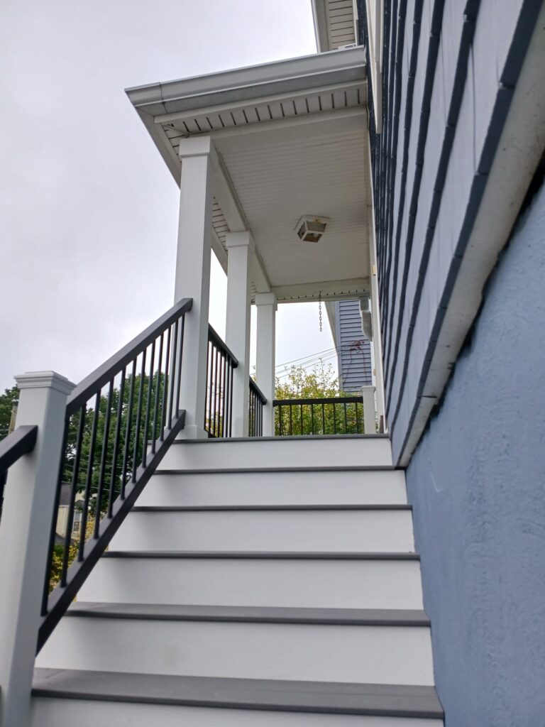 Newly renovated outdoor staircase with white risers and gray treads leading to a front porch of a two-story house in Brighton, MA. The stairs feature sleek black metal railings with white posts, complementing the modern look of the gray-blue siding and white trim. Overhead, a small porch roof with square columns and a ceiling light fixture provides shelter at the entryway