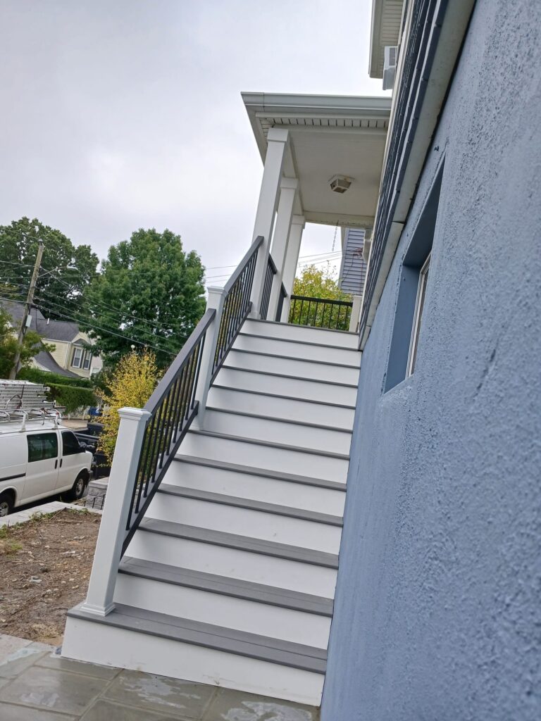 Finished porch stairs by Monroy's Carpenters Inc in Brighton MA: Gray composite stairs with black metal railings leading to a white porch on a blue house, ideal for outdoor access in Greater Boston homes.